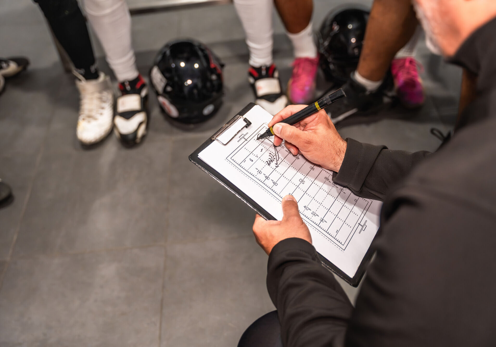 Football coach sketching a new play on a clipboard in the locker room while discussing strategy with players, focused planning and leadership before the game
